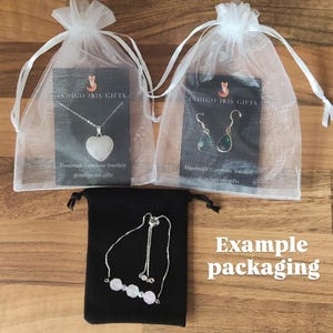 May include: Example packaging for handmade gemstone jewelry. Three white organza bags with black cards that say "Indigo Iris Gifts" and feature a necklace with a heart-shaped gemstone, a pair of earrings with green gemstones, and a bracelet with pink gemstones. The bags are on a wooden surface.