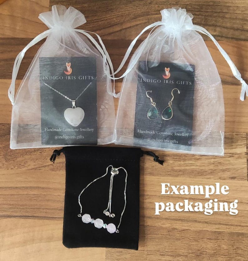 May include: Example packaging for handmade gemstone jewelry. Three white organza bags with black cards that say "Indigo Iris Gifts" and feature a necklace with a heart-shaped gemstone, a pair of earrings with green gemstones, and a bracelet with pink gemstones. The bags are on a wooden surface.