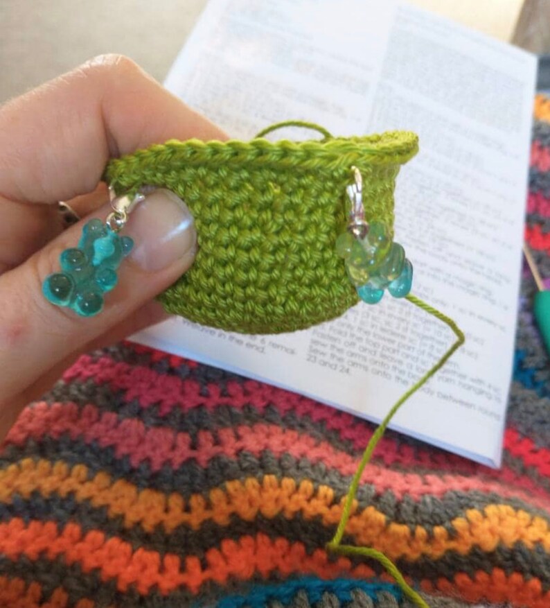 Puede incluir: Una taza de ganchillo verde con dos abalorios de osito de goma turquesa unidos al borde. La taza est&aacute; sobre una manta de ganchillo de rayas multicolores.