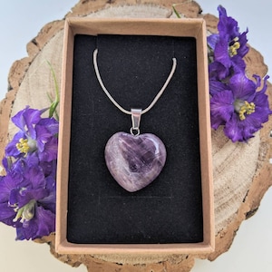 May include: A silver chain necklace with a heart-shaped amethyst pendant. The pendant is a deep purple color and has a natural, polished finish. The necklace is displayed in a brown box with a black velvet lining.