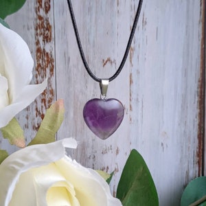 May include: A purple amethyst heart pendant on a black cord necklace. The pendant is hanging against a white wooden background.