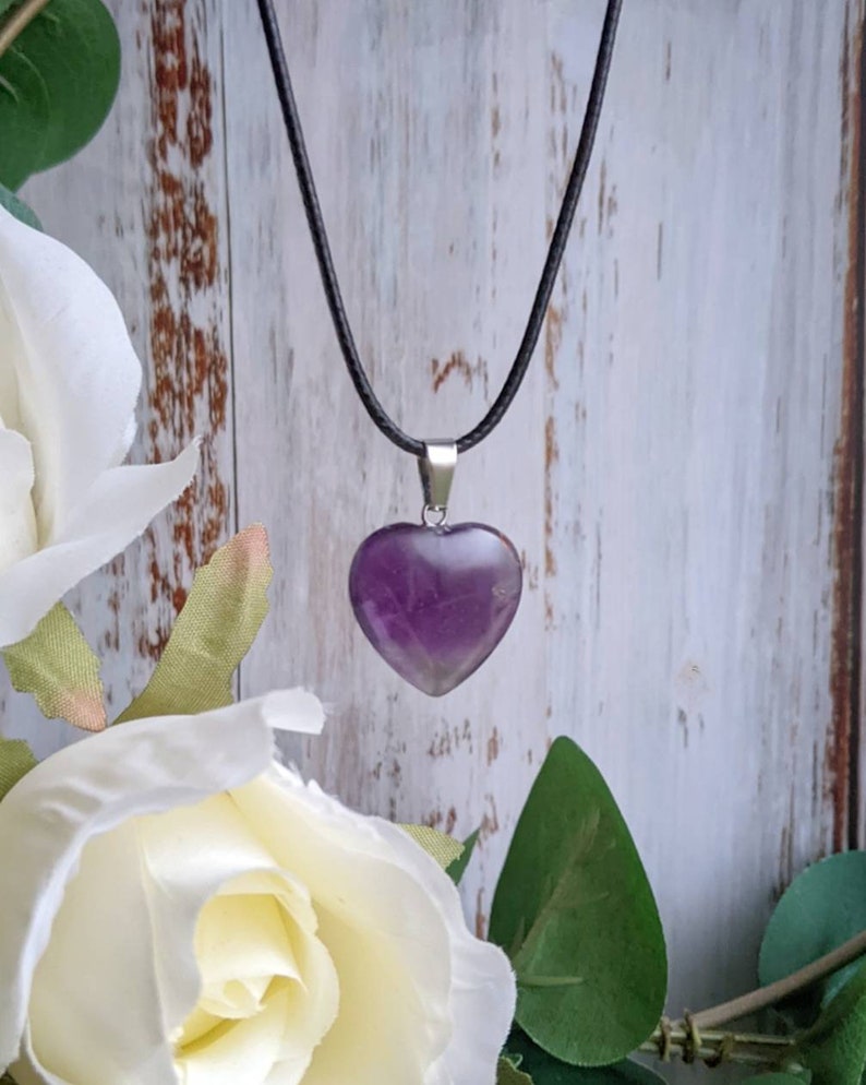 May include: A purple amethyst heart pendant on a black cord necklace. The pendant is hanging against a white wooden background.