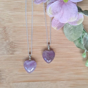 May include: Two silver chains with amethyst heart pendants. The hearts are a light purple color and are on a wooden background.