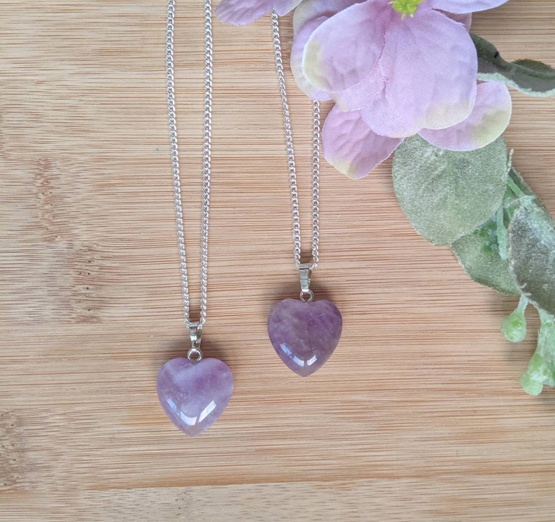 May include: Two silver chains with amethyst heart pendants. The hearts are a light purple color and are on a wooden background.