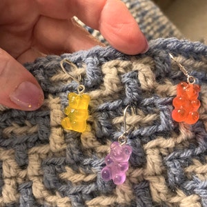 Puede incluir: Tres pendientes con un encanto de osito de goma en una tela de crochet azul y beige. Los pendientes son amarillos, naranjas y morados.
