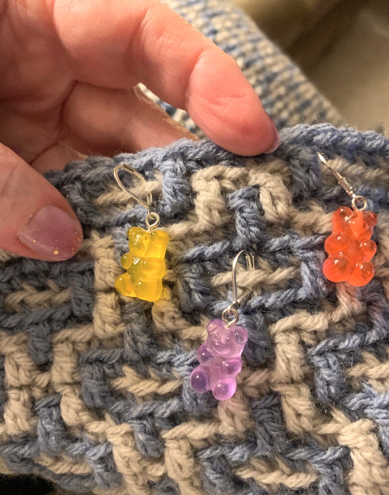 Puede incluir: Tres pendientes con un encanto de osito de goma en una tela de crochet azul y beige. Los pendientes son amarillos, naranjas y morados.