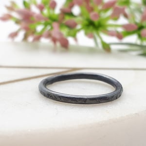 Könnte beinhalten: Ein dünner schwarzer Ring mit einer strukturierten Oberfläche. Der Ring liegt auf einer weißen Marmorfläche.