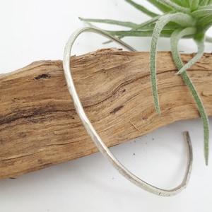 Könnte beinhalten: Ein silberfarbenes, gehämmertes Metallarmband mit offenem Design. Das Armband befindet sich vor einem natürlichen Holzgrund mit einer grünen Pflanze. Das Armband hat eine strukturierte Oberfläche und einen schlichten, eleganten Stil.