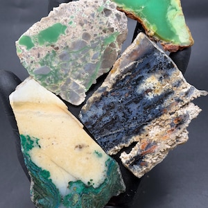May include: A collection of raw, polished gemstones in various colors. The stones include shades of green, blue, white, and brown. The textures vary from smooth to rough, showcasing natural patterns and mineral formations. The gemstones are displayed against a dark background.