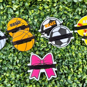 Könnte beinhalten: Eine Sammlung von fünf runden Kabelorganisatoren mit Sportmotiven und einem pinkfarbenen, schleifenförmigen Kabelorganisator. Jeder Sportartikel zeigt eine Ballgrafik, darunter Volleyball, Basketball, Fußball und Softball, mit dem Wort NAME darauf.