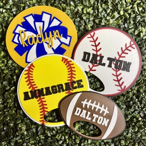 Könnte beinhalten: Vier personalisierte Flaschenöffner im Sportdesign. Einer in Gelb mit blau-weißem Pompon-Motiv und der Aufschrift 'Radyn', ein weißer Baseball mit roten Nähten und 'DALTON', ein gelber Softball mit 'ANNAGRACE' und ein brauner Football mit 'DALTON'.