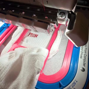 K&ouml;nnte beinhalten: Eine Stickmaschine mit einem wei&szlig;en Stoffst&uuml;ck in einem rosa Rahmen. Die Maschine hat eine Nadelstange mit nummerierten Schlitzen und ein wei&szlig;es Geh&auml;use. Auf dem Stoff ist der rote Sticktext PION zu sehen. Die Basis zeigt U.S. PAT. NOS. 7,607,399 & 7,918,169.