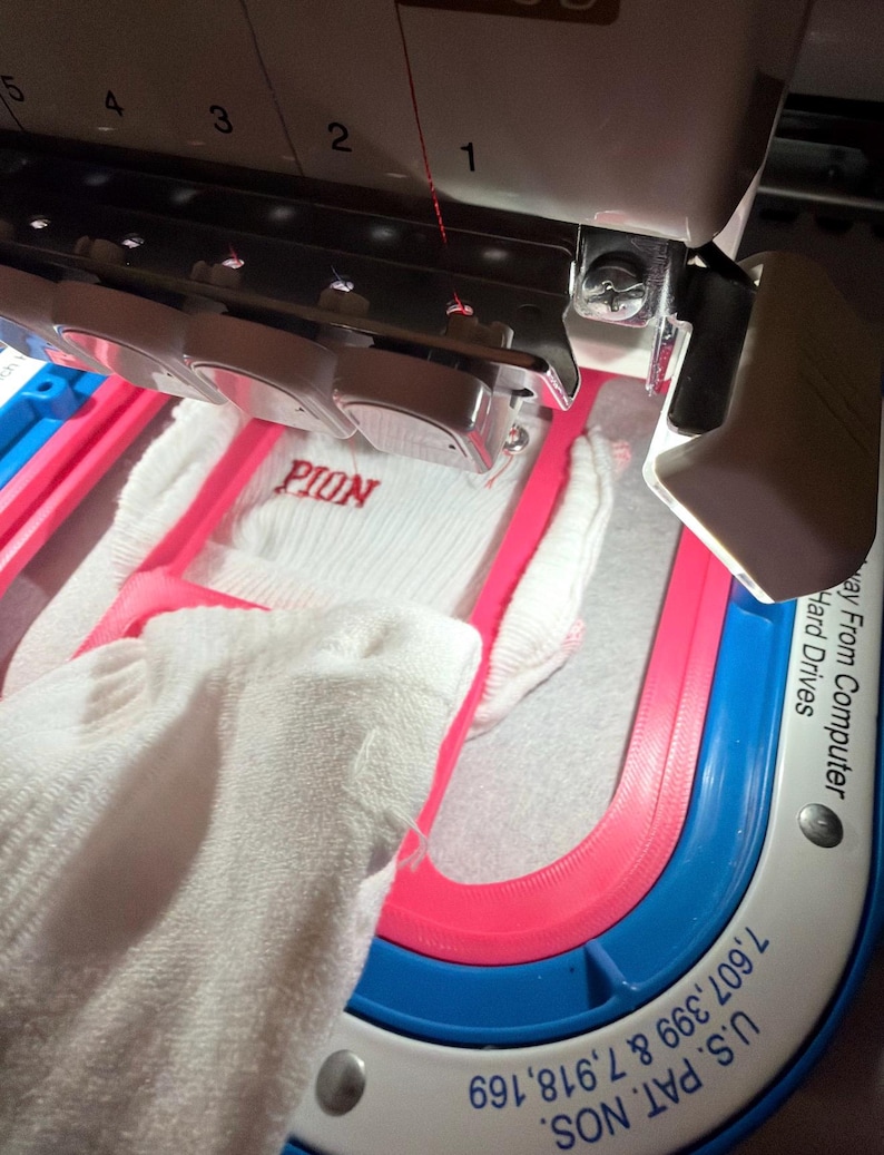 K&ouml;nnte beinhalten: Eine Stickmaschine mit einem wei&szlig;en Stoffst&uuml;ck in einem rosa Rahmen. Die Maschine hat eine Nadelstange mit nummerierten Schlitzen und ein wei&szlig;es Geh&auml;use. Auf dem Stoff ist der rote Sticktext PION zu sehen. Die Basis zeigt U.S. PAT. NOS. 7,607,399 & 7,918,169.