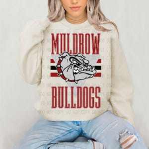 May include: Cream sweatshirt with the text "Muldrow Bulldogs" in red. The design includes a bulldog graphic with a red and black border. The sweatshirt is worn by a person wearing blue jeans.