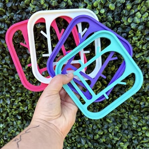 Peut inclure: Un ensemble de quatre cadres carrés en plastique aux couleurs rose vif, blanc, violet et turquoise. Chaque cadre présente une découpe centrale rappelant un motif de branche d'arbre stylisée, avec des coins arrondis et une surface lisse.
