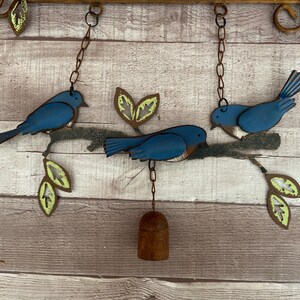May include: A metal wind chime with three blue birds perched on a branch with green leaves. The birds are hanging from chains and a bell hangs from the bottom of the chime.