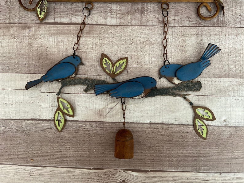 May include: A metal wind chime with three blue birds perched on a branch with green leaves. The birds are hanging from chains and a bell hangs from the bottom of the chime.