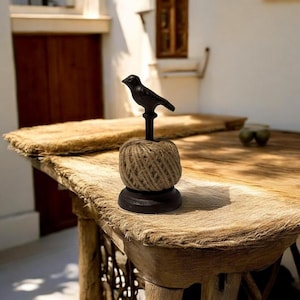 May include: A decorative twine dispenser featuring a black bird figurine atop a twine ball. The dispenser sits on a dark brown base. The twine ball is made of natural-colored jute twine. The dispenser is placed on a wooden table.