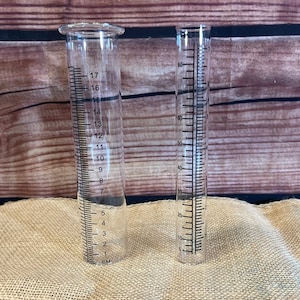 May include: Two clear glass rain gauges with black markings. The gauge on the left measures in centimeters up to 17 cm. The gauge on the right measures in millimeters up to 80 mm.