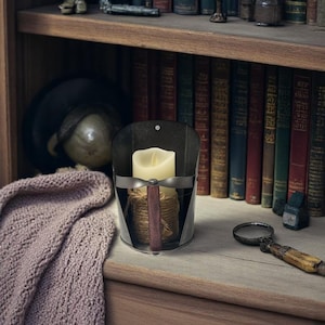 May include: A black metal candle holder with a cream-colored candle, rope, and a wooden handle. The candle holder is on a wooden shelf with books and a magnifying glass. A knitted scarf is draped on the shelf.