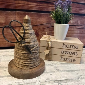 May include: A wooden spool wrapped in twine with a pair of scissors sticking out of the top. The spool is sitting on a wooden base. The background is a wooden wall with a potted plant and a stack of books that say "home sweet home".