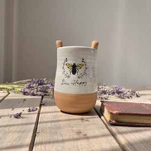 May include: A ceramic vase with a cream-colored top and a tan base. The vase features a black and yellow bee design with the words "Bee Happy" written below. Two small tan handles are on the top. The vase is on a wooden surface.