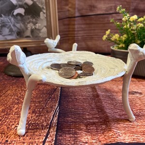 May include: A white, bird-shaped trinket dish with a textured surface, holding several coins. The dish is supported by three bird-like legs. A framed photo and potted plant are in the background.