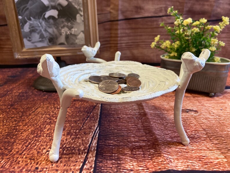 May include: A white, bird-shaped trinket dish with a textured surface, holding several coins. The dish is supported by three bird-like legs. A framed photo and potted plant are in the background.