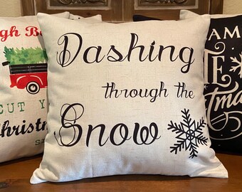 May include: Off-white square pillow with the phrase "Dashing through the Snow" in black script, accompanied by a black snowflake. Other pillows feature Christmas-themed text and graphics, including a red truck carrying a Christmas tree.