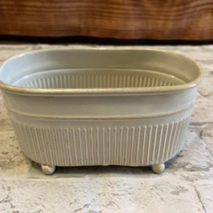 May include: A rectangular, off-white ceramic planter with a ribbed exterior and rounded corners. The planter sits on three small, rounded feet. The rim has a subtle, light-colored trim. This decorative piece is suitable for small plants or as a decorative accent.