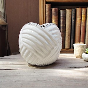 May include: A white ceramic decorative knot-shaped vase with a textured surface and black speckling. The vase is on a wooden table, with a bookshelf and other small planters in the background.