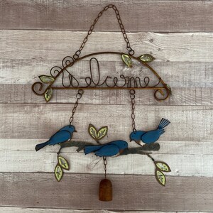 May include: A metal welcome sign with blue birds and leaves hanging from it. The sign is rusty and has a rustic look.