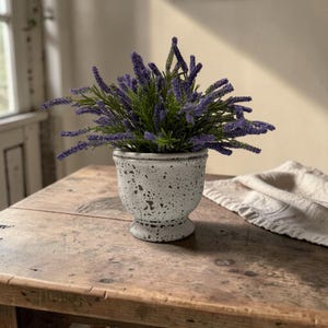 May include: A small, speckled, off-white ceramic pot with a pedestal base, filled with faux lavender. The lavender has purple flower spikes and green leaves. The pot sits on a rustic, wooden table.