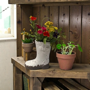 May include: A white boot-shaped planter filled with colorful flowers, including red, yellow, and purple blooms. A small succulent and a potted plant sit nearby on a rustic wooden shelf. The boot planter has a speckled design and a brown base.