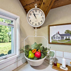 May include: A white, round wall clock with black numerals and hands hangs above a white metal basket filled with fruit and greenery. The clock reads "GRAND HOTEL 1870". A framed picture of a house is on the wall.