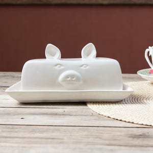 May include: White ceramic butter dish shaped like a pig with a lid and a separate base. The pig has two ears and a snout.