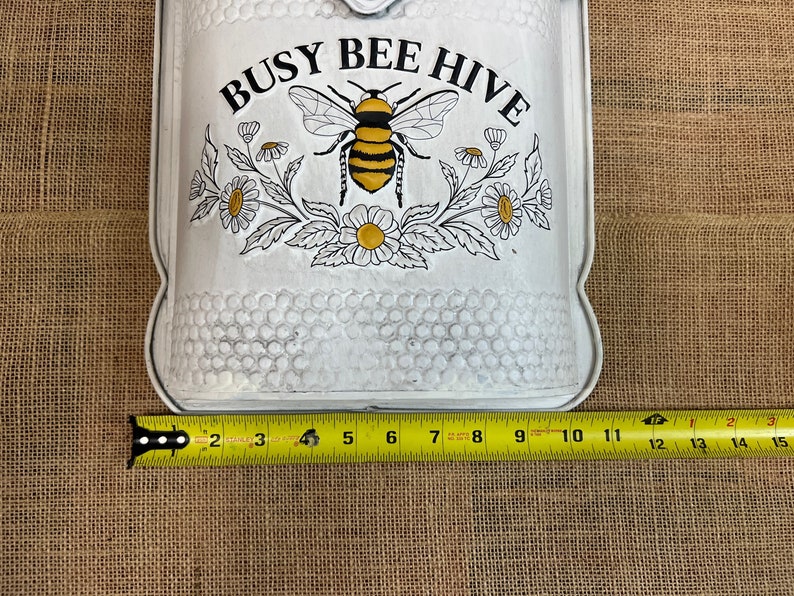May include: White metal mailbox with a black and yellow bee and flowers. The mailbox has the words "Busy Bee Hive" printed on it. The mailbox is 10 inches wide.
