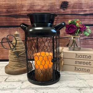 May include: Black metal candle holder with a wire cage design. The candle holder is shaped like a milk jug and has a warm orange candle inside. The candle holder is sitting on a table with a wooden sign that says "home sweet home".