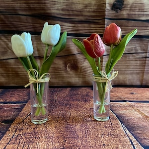 May include: Two small glass vases with a simple design, one with white tulips and the other with red tulips. Both vases are tied with a natural twine bow.