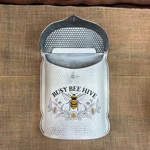 May include: White metal mailbox with a bee and flower design. The mailbox has a flap that opens to reveal a slot for mail. The mailbox has the text "Busy Bee Hive" on the front.