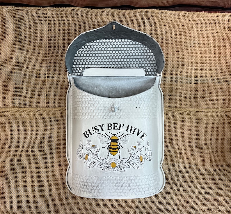 May include: White metal mailbox with a bee and flower design. The mailbox has a flap that opens to reveal a slot for mail. The mailbox has the text "Busy Bee Hive" on the front.