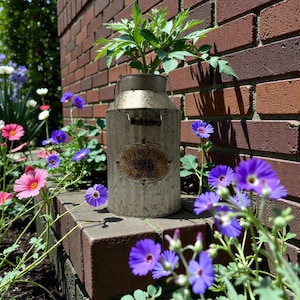 May include: A galvanized metal milk can planter with a handle, holding a green plant. The planter has a decorative oval label that reads "FLOWERS PLANTS". The planter sits on a brick ledge, surrounded by colorful flowers.