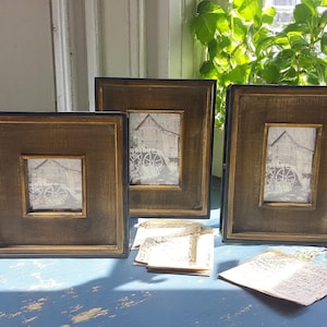 May include: Three square picture frames with a rustic, distressed wood finish. Each frame has a black border and a gold inner frame. The pictures inside are black and white images of a building with a water wheel. The frames are on a blue surface.