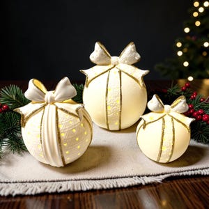 May include: Three spherical, cream-colored ceramic ornaments shaped like wrapped gifts, each topped with a bow and accented with gold glitter trim. The surfaces feature intricate cut-out patterns that emit a warm, soft glow from within.
