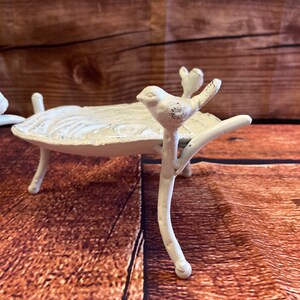 May include: A white, cast-iron bird bath with three legs and a leaf-shaped basin. Two small birds perch on the rim, and another sits on a branch extending from the base. The bird bath has a distressed, antique finish.