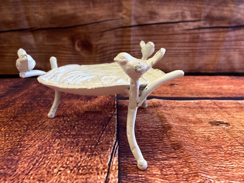 May include: A white, cast-iron bird bath with three legs and a leaf-shaped basin. Two small birds perch on the rim, and another sits on a branch extending from the base. The bird bath has a distressed, antique finish.