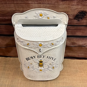 May include: White metal mailbox with a busy bee hive design. The mailbox has a floral pattern with daisies and a bee with yellow and black stripes. The mailbox has a slot for mail and a small opening for the mail to be retrieved.