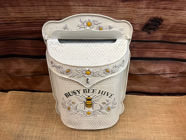 May include: White metal mailbox with a busy bee hive design. The mailbox has a floral pattern with daisies and a bee with yellow and black stripes. The mailbox has a slot for mail and a small opening for the mail to be retrieved.