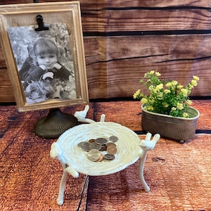 May include: A decorative display featuring a framed 5x7 inch black and white photo, a white bird-shaped dish holding coins, and a small metal planter with greenery. The frame has a wooden appearance, and the dish has a distressed finish.