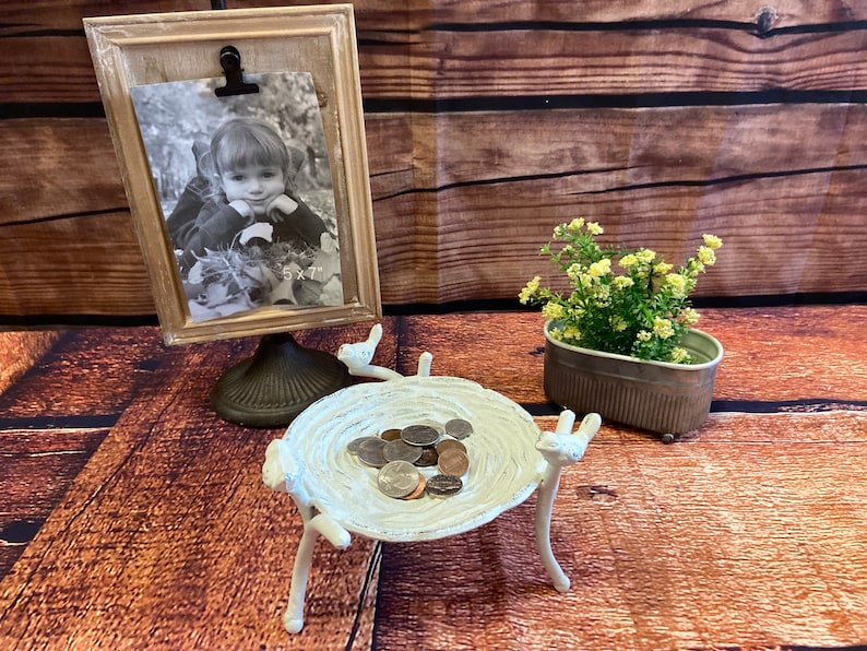 May include: A decorative display featuring a framed 5x7 inch black and white photo, a white bird-shaped dish holding coins, and a small metal planter with greenery. The frame has a wooden appearance, and the dish has a distressed finish.